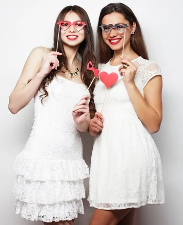 Two women in white dresses holding photo booth props, including red glasses and a heart sign, smiling at the camera.