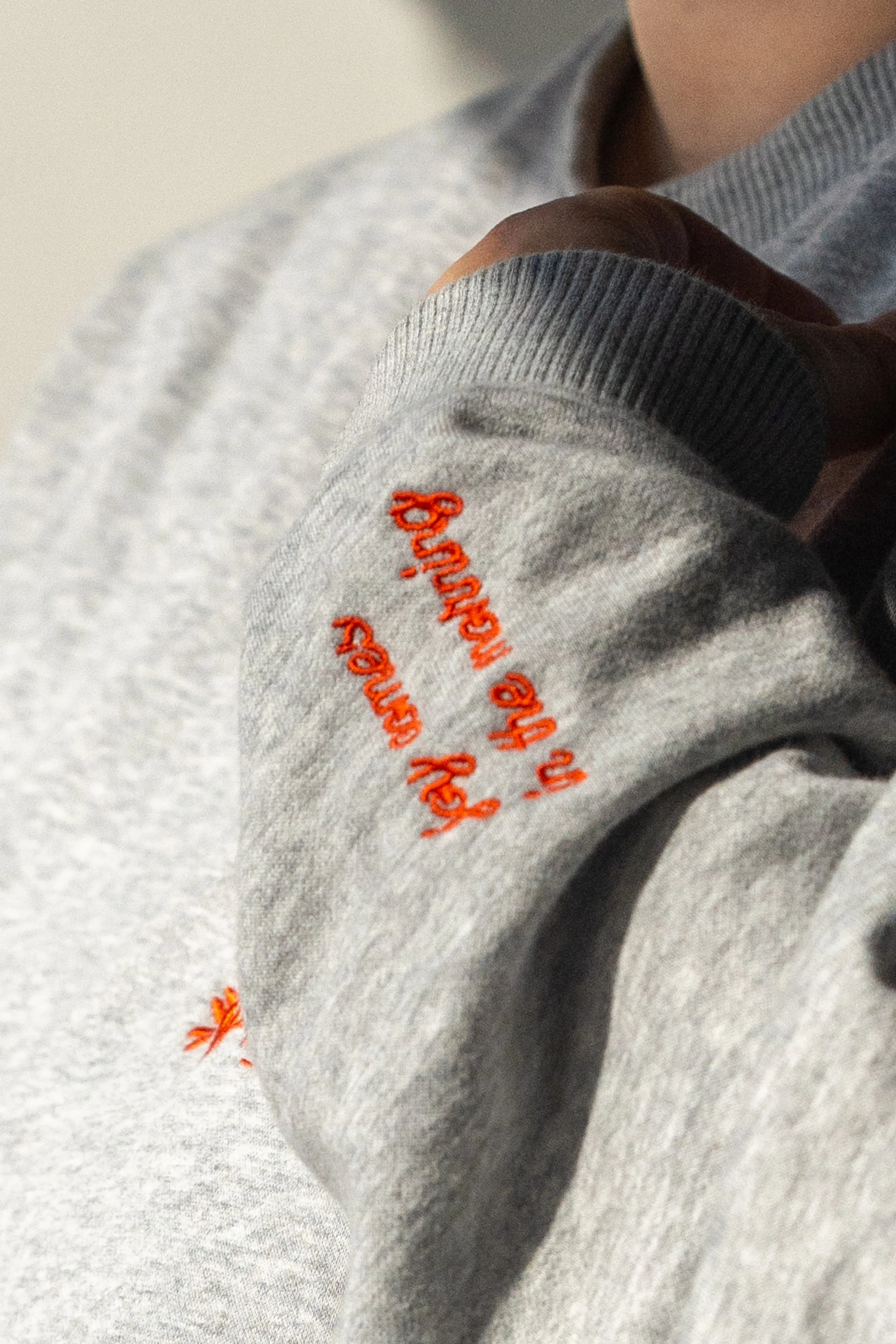 グレーのスウェットの左袖に小さく赤い刺繍で「Joy comes in the morning」と書かれている