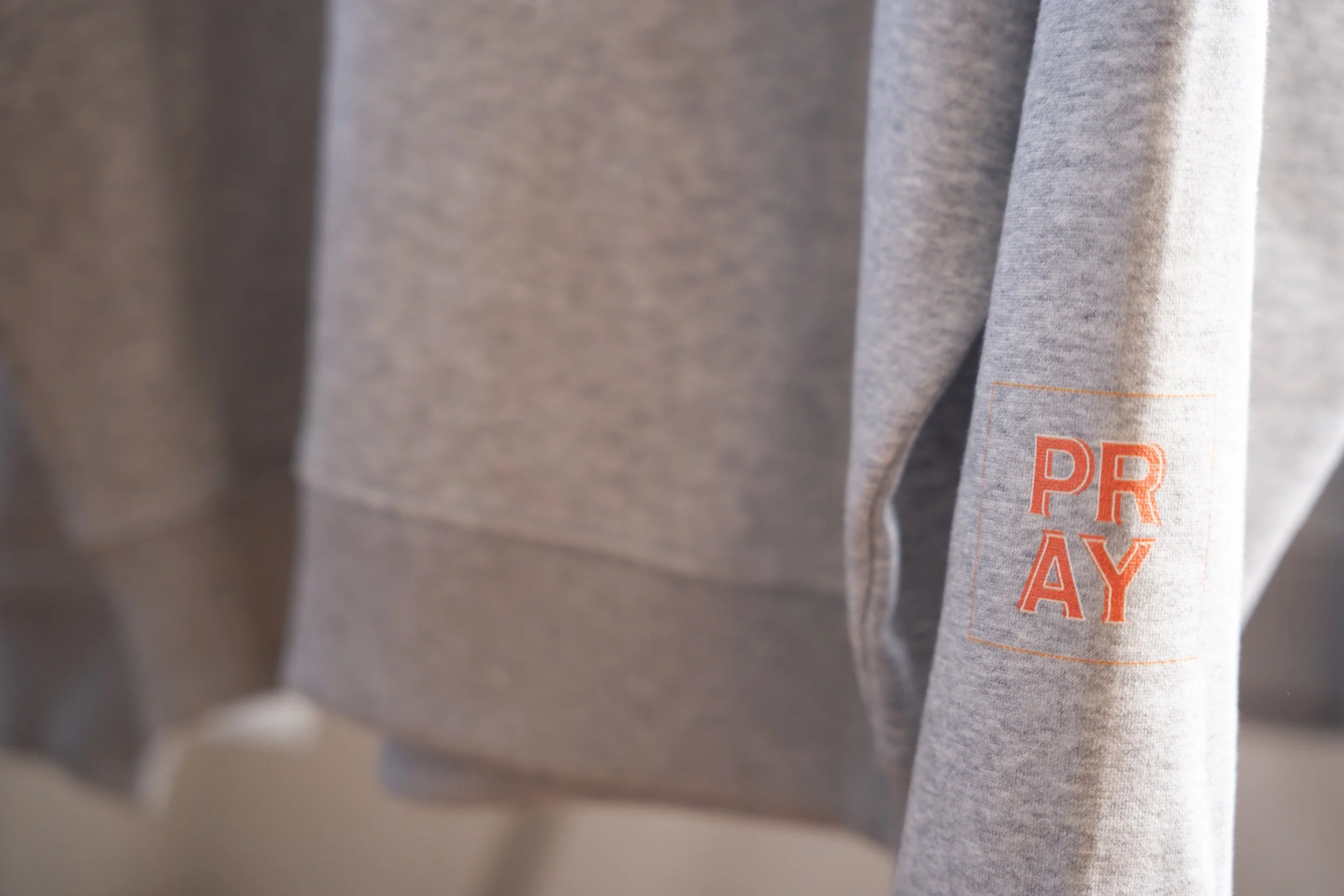 灰色のスウェットシャツの袖にオレンジ色の文字で「PRAY」と書かれている