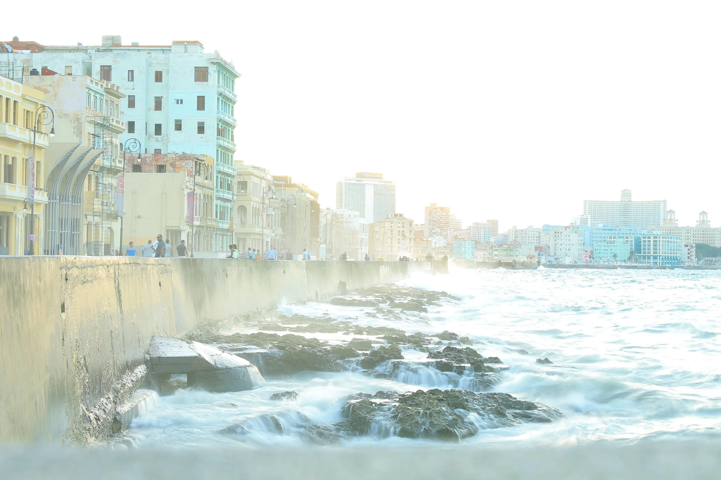 海辺の遊歩道と波打つ岩礁、背景にカラフルな古い建物群と高層ビル群、夕日が差し込んでいるキューバ、ハバナの川沿い都市景観