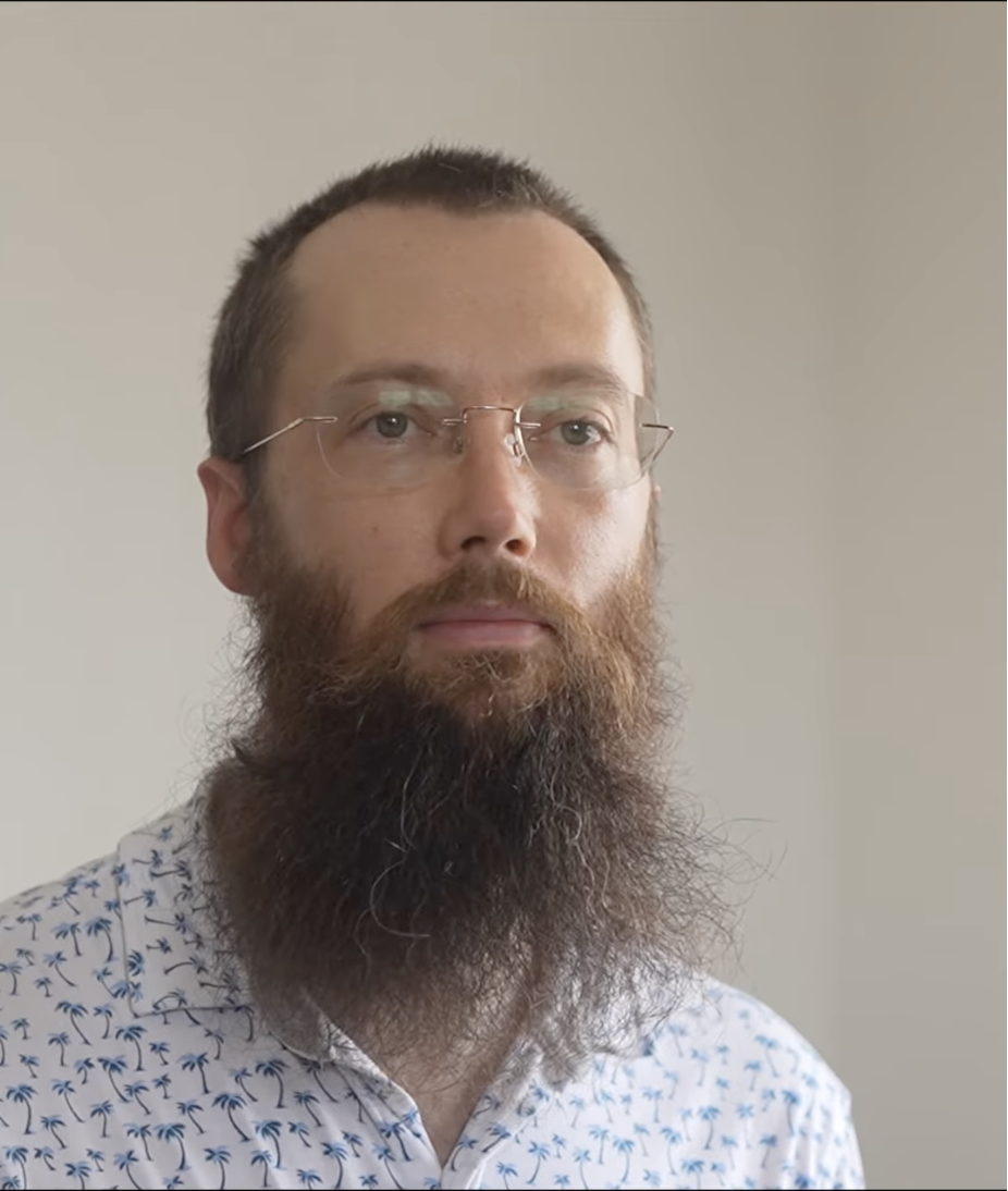 メガネをかけた男性のポートレート写真。白いシャツを着ていて、背景は単色の壁。