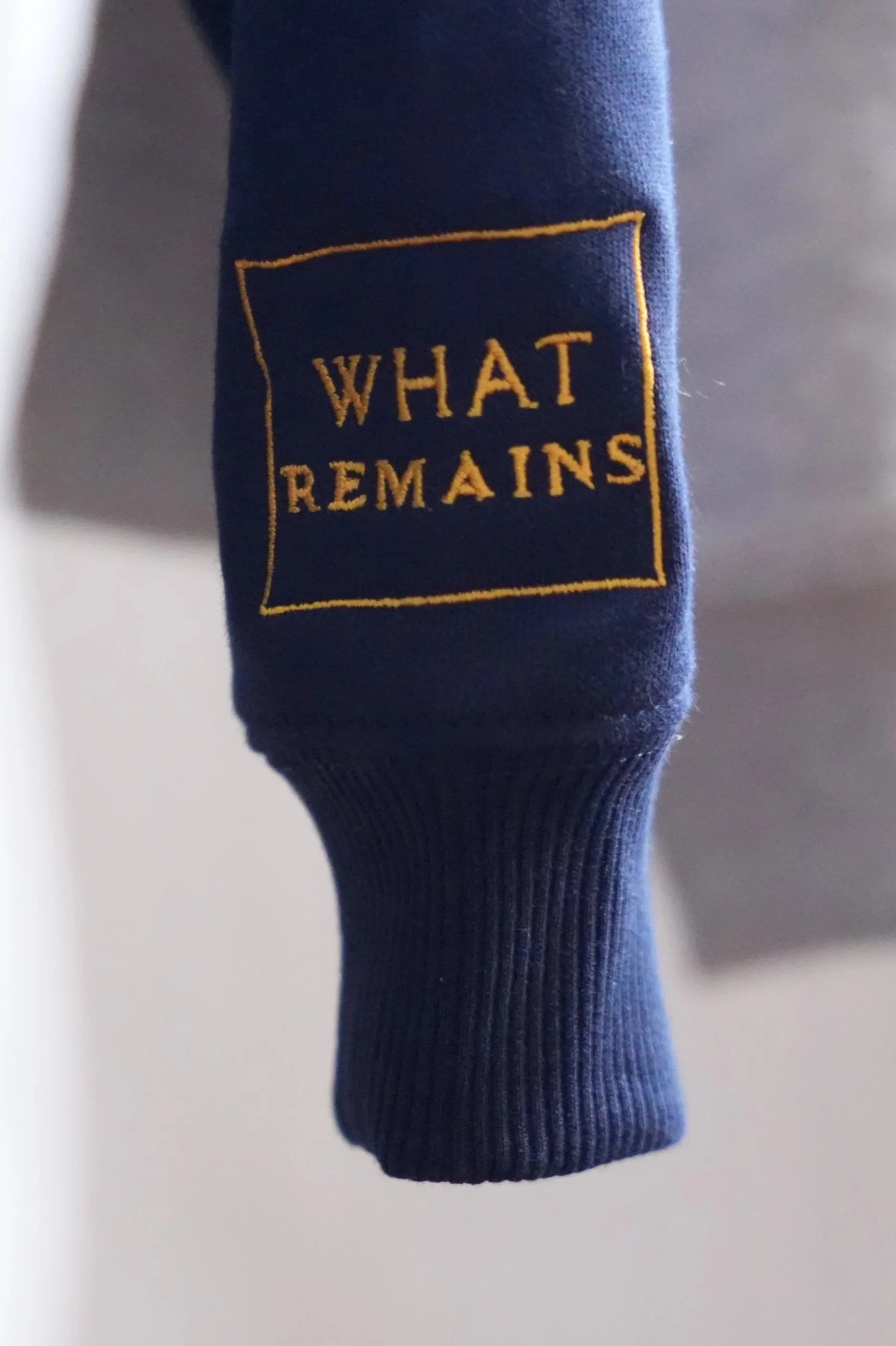 紺色の袖口に黄色の刺繍で「WHAT REMAINS」と書かれたデザインの衣服