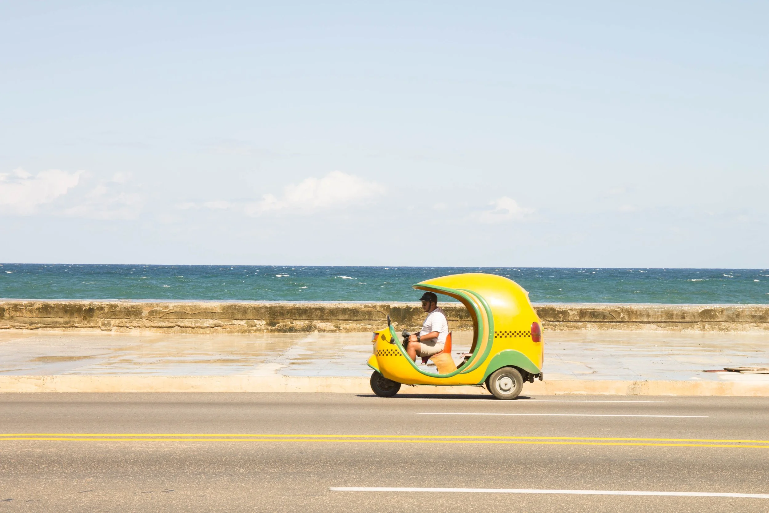 キューバ、ハバナ海沿いの道路を走る黄色と緑のアイスクリーム型の三輪タクシーを運転するタクシードライバー男性