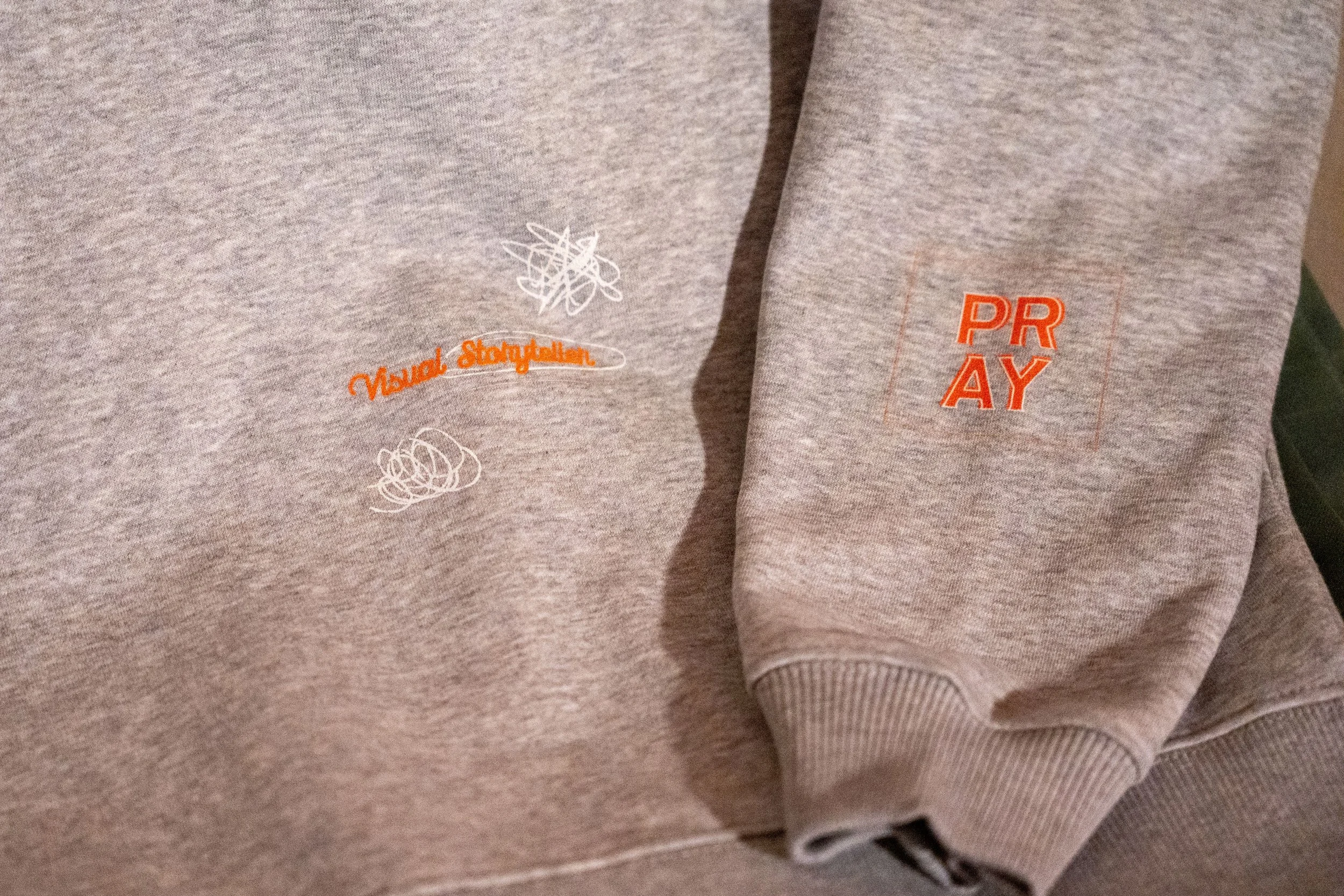 グレーのスウェットにオレンジと白で「Visual Storyteller」「PRAY」とプリントされてある
