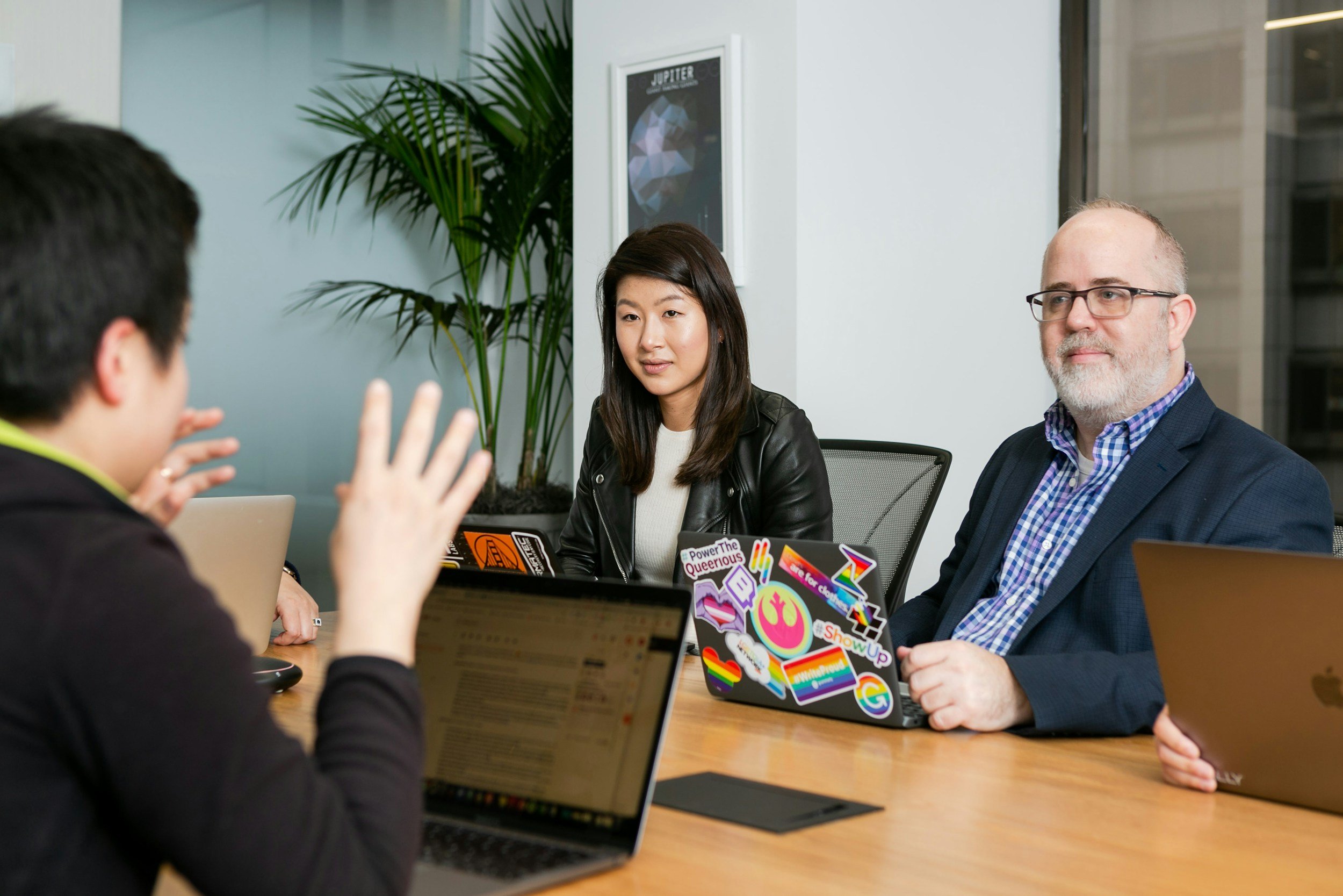 Four people in a meeting room having a discussion. One person is gesturing with hands, while two others are listening. All have laptops in front of them, with the woman and the man on the right having decorated laptops. The room has a plant and a framed picture of Jupiter on the wall.