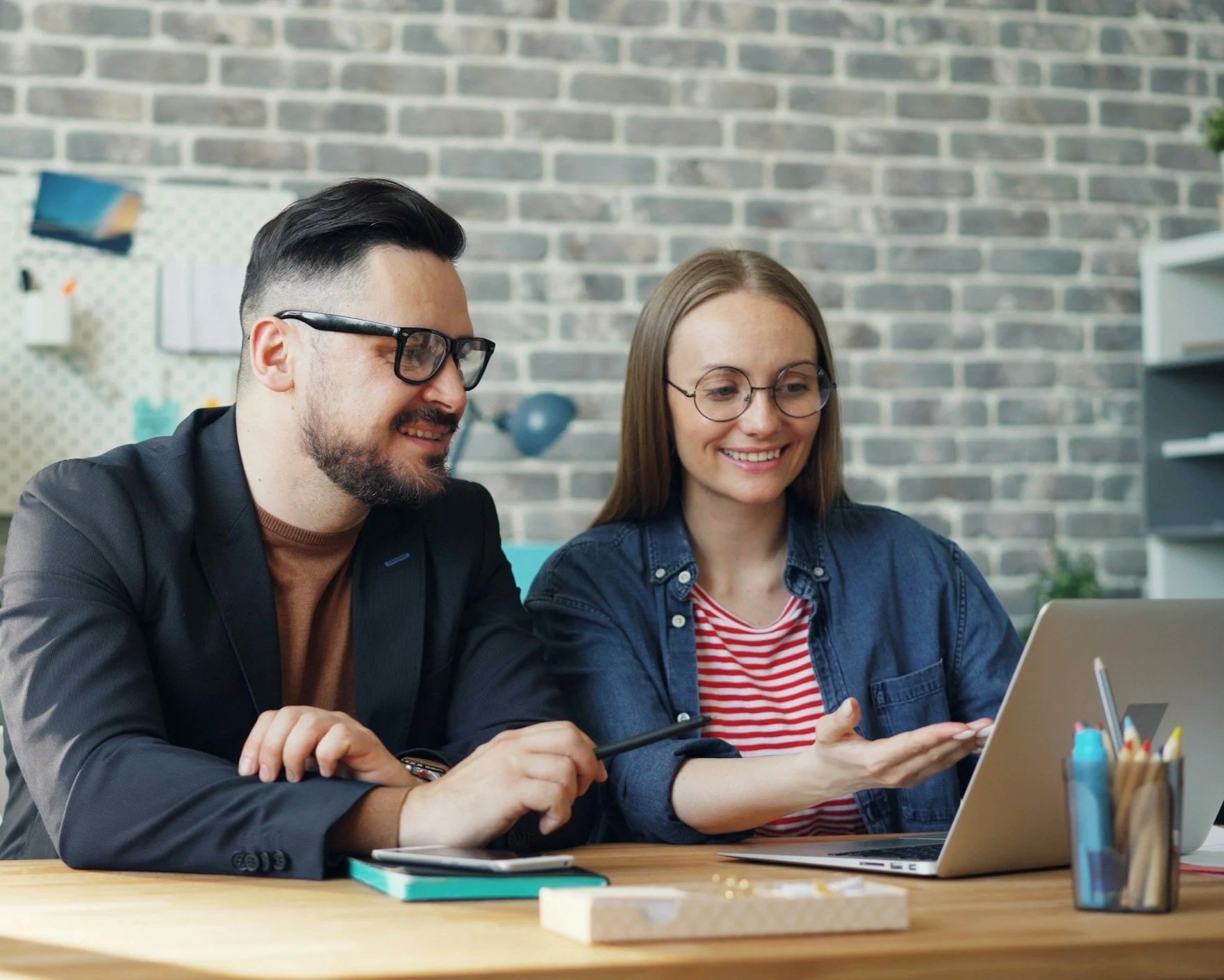 Two people, a man and a woman, sitting at a desk looking at a laptop together and smiling, in a modern office with a brick wall background.