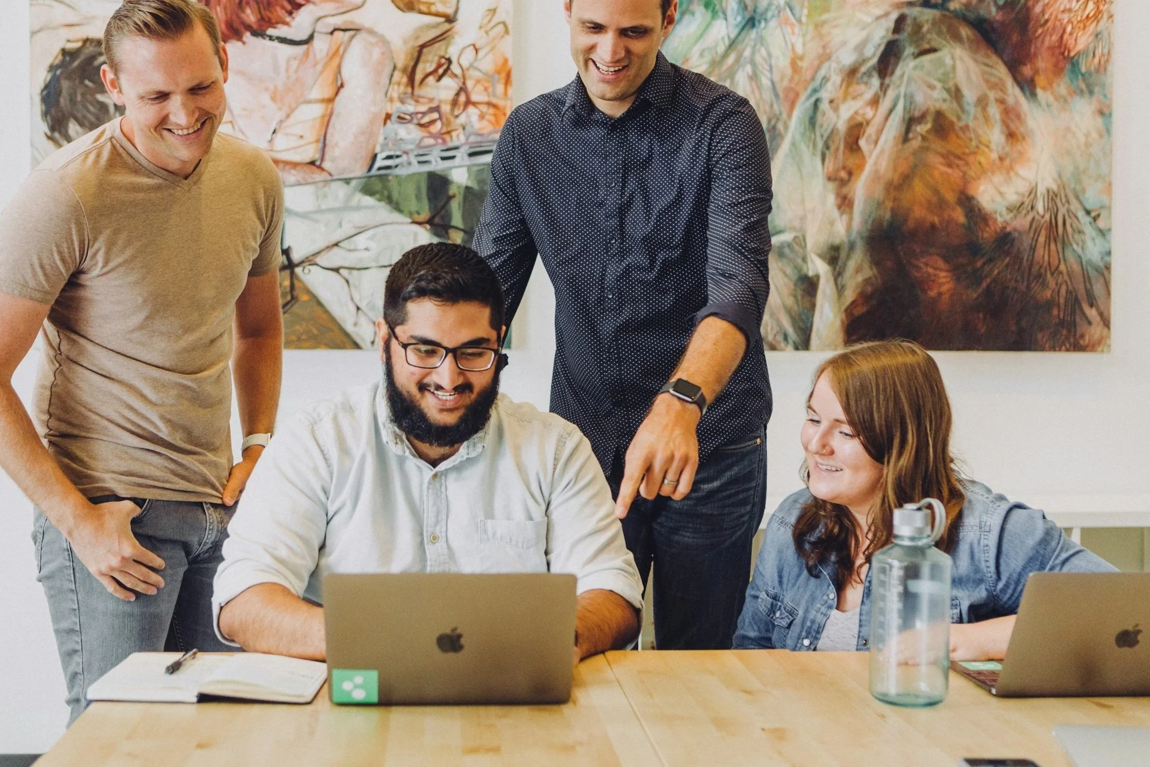 Four people gathered around a table, looking at a laptop, smiling, with two of them standing and pointing at the laptop screen, and artwork hanging on the wall behind them.