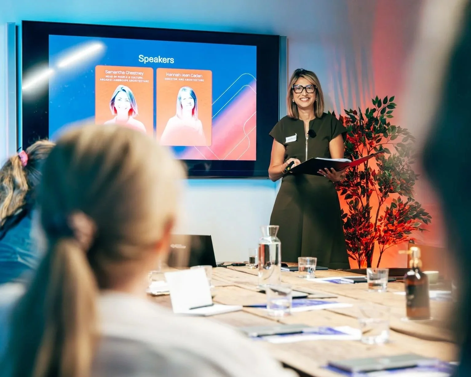 A woman presenter standing in front of a large screen with speaker profiles, holding a notebook, during a conference or meeting. Attendees are seated at a table with water glasses, notebooks, and a laptop.