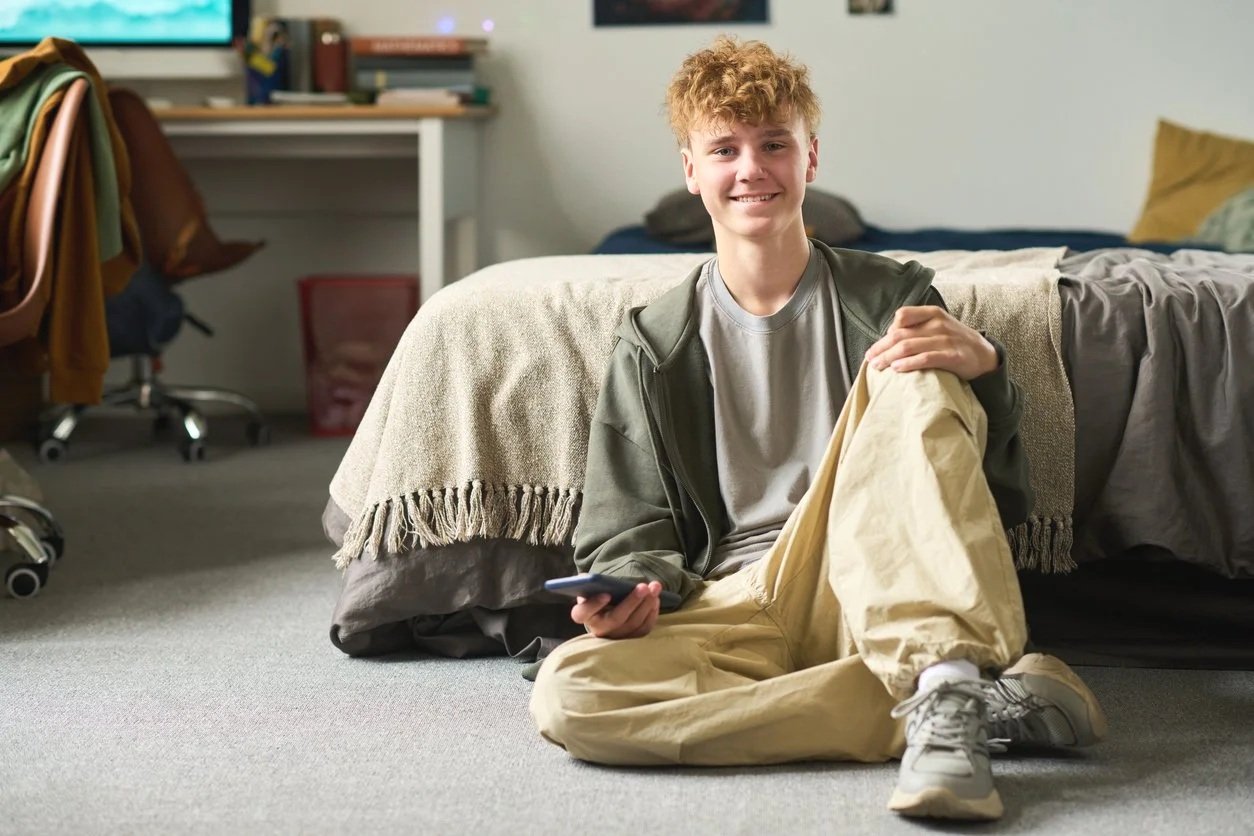A young man sitting on the floor next to a bed, smiling at the camera, holding a TV remote in his right hand, and wearing casual clothes including a gray t-shirt, beige pants, and sneakers.