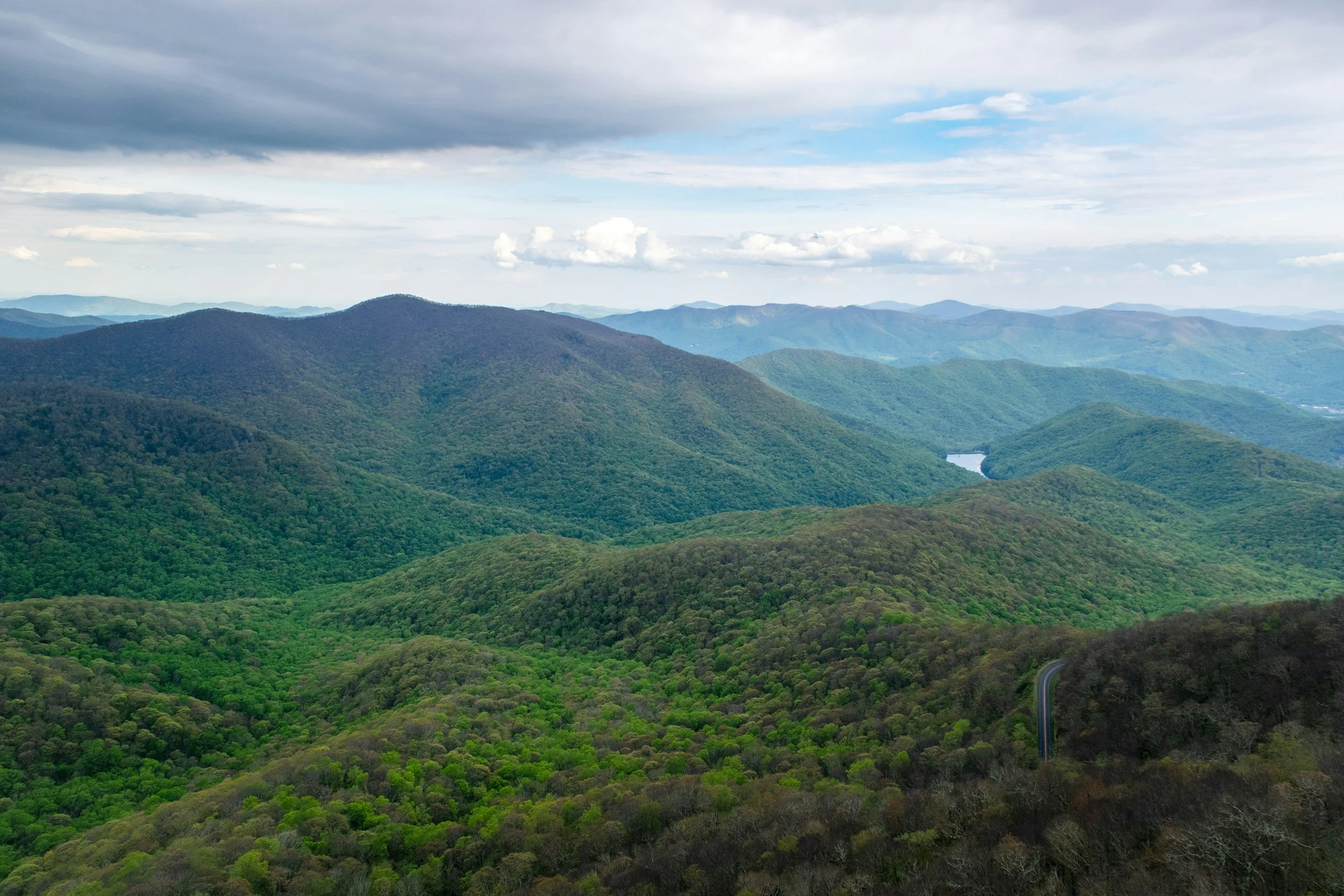 A panoramic view of green forested mountains under a partly cloudy sky, with a winding road visible in the lower right corner.