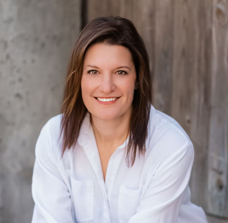 Headshot of a woman with shoulder-length brown hair, smiling, wearing a white blouse, standing outdoors with a wooden fence in the background.