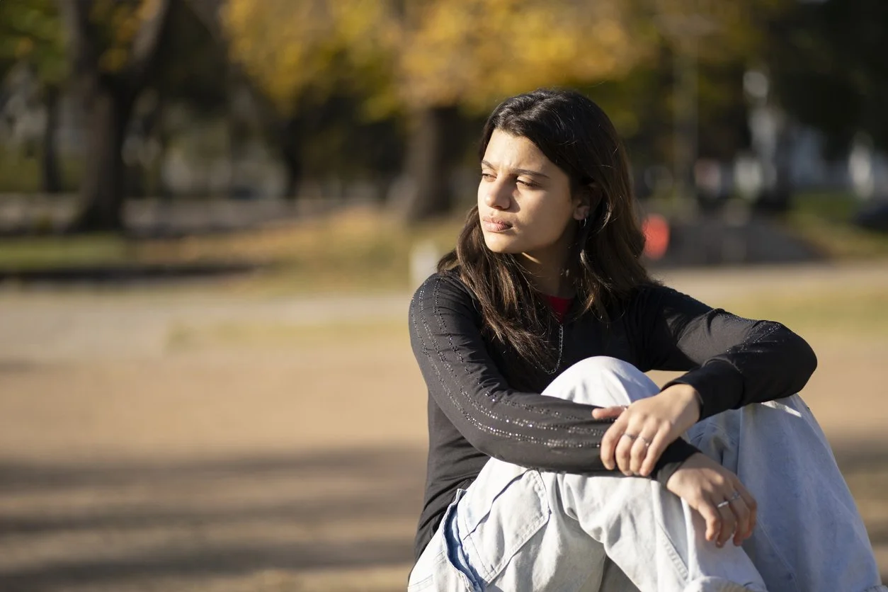 A young woman sitting outdoors on a sunny day, wearing a black long-sleeve shirt and white pants, looking contemplative with her eyes closed.