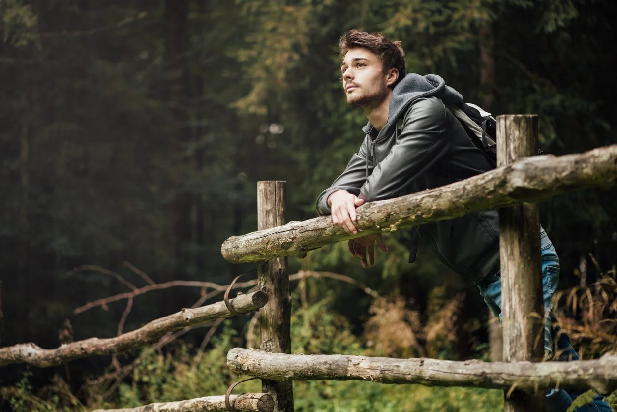 A young man with dark hair and facial hair leaning on a rustic wooden fence in a forest, looking thoughtfully into the distance.