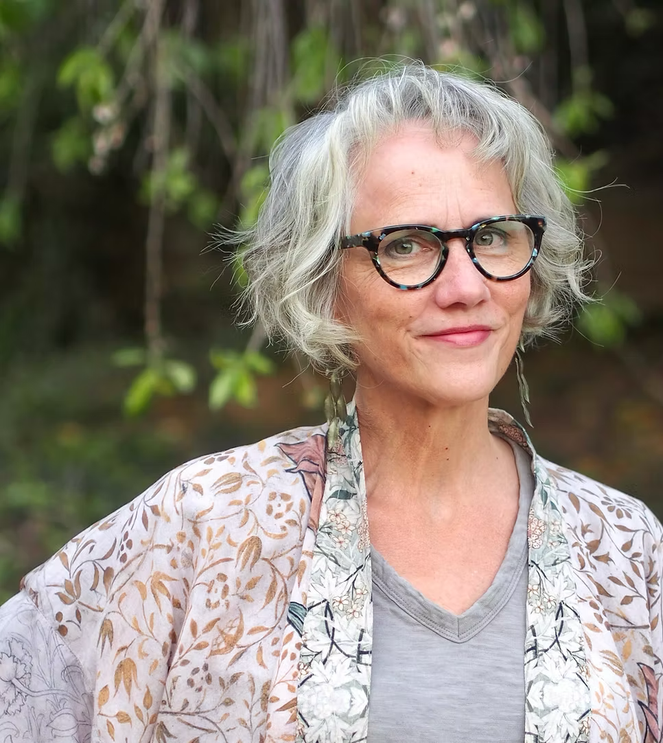 A woman with short, curly gray hair wearing tortoiseshell glasses and a light patterned shawl over a gray t-shirt outdoors with greenery in the background.