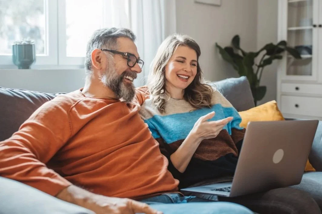 A middle-aged man with glasses and a beard, wearing an orange long-sleeve shirt, and a young woman with curly hair, wearing a striped sweater, sitting together on a sofa looking at a laptop and smiling.