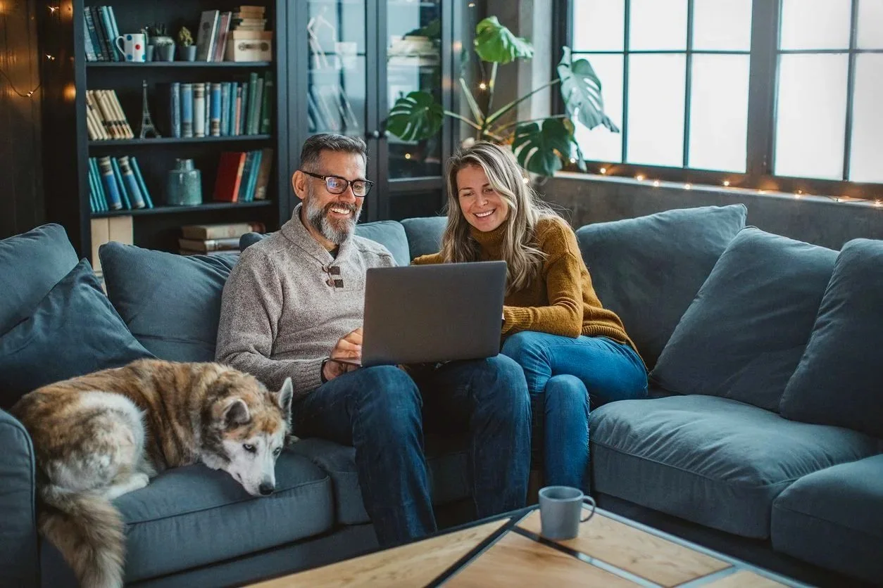 A man and woman sitting on a gray sofa in a cozy living room, looking at a laptop and smiling, with a dog lying beside them. The room has large windows, a bookshelf, and a large green plant.