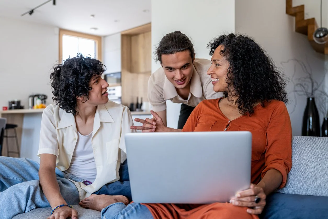 Three people having a conversation in a modern kitchen, with one person sitting on a sofa using a laptop, another sitting nearby, and a third standing close to them, engaging in the discussion.