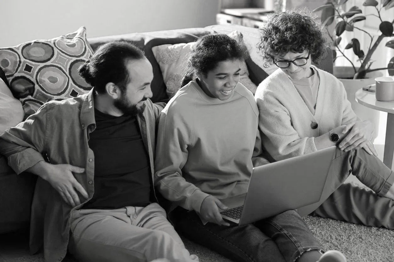 Three friends sitting on the floor in a living room, looking at a laptop and smiling.