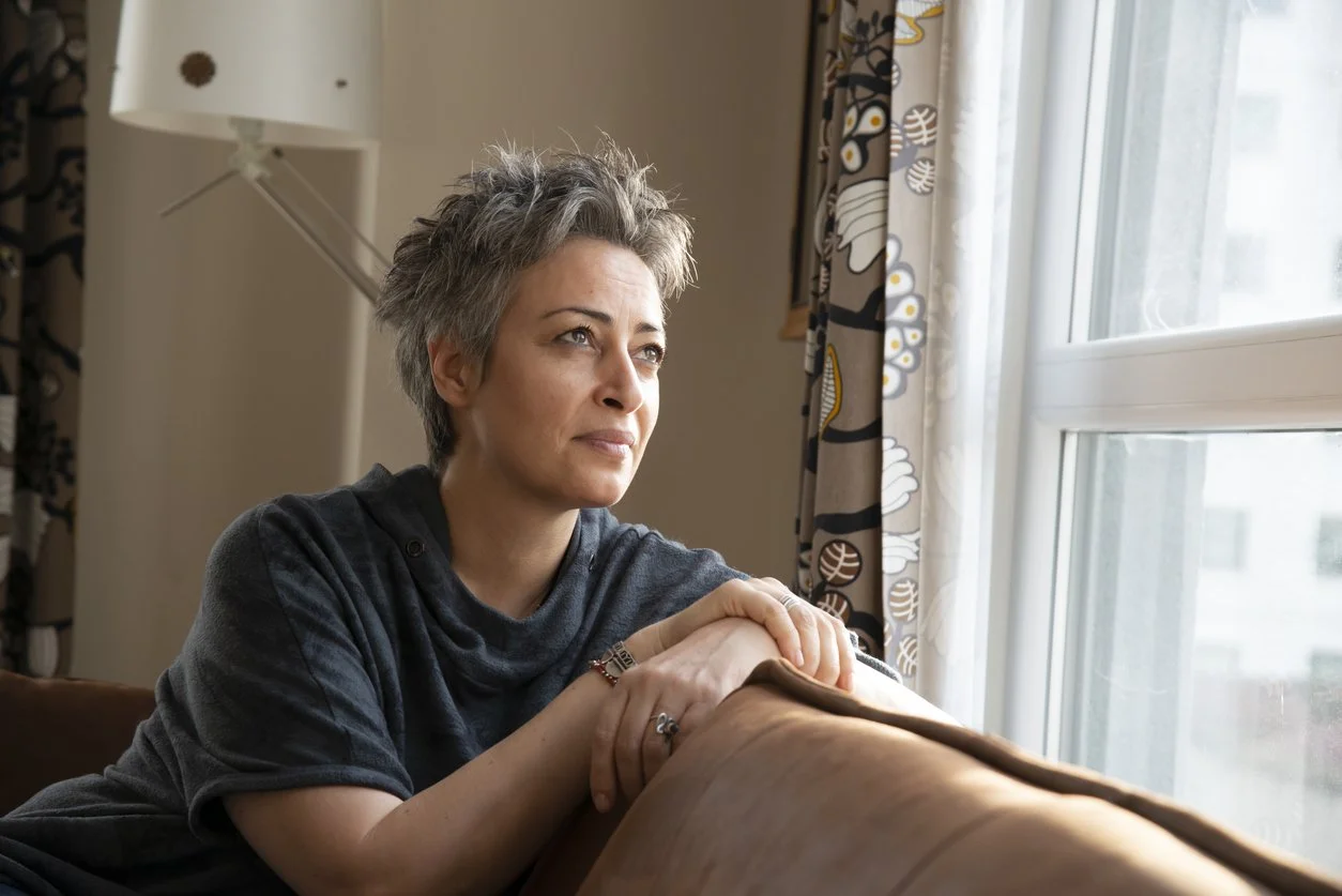 Woman with short gray hair looking pensively out the window while leaning on a brown couch armrest in a living room.