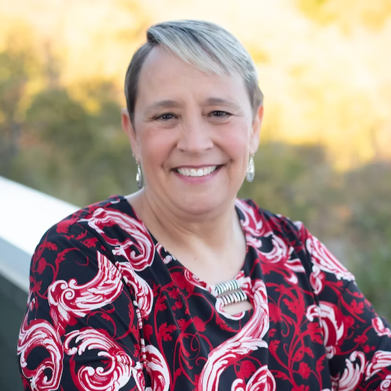 A woman with short blonde hair, wearing a black and red patterned blouse and silver jewelry, smiling outdoors with a blurred background of trees and a river during daytime.