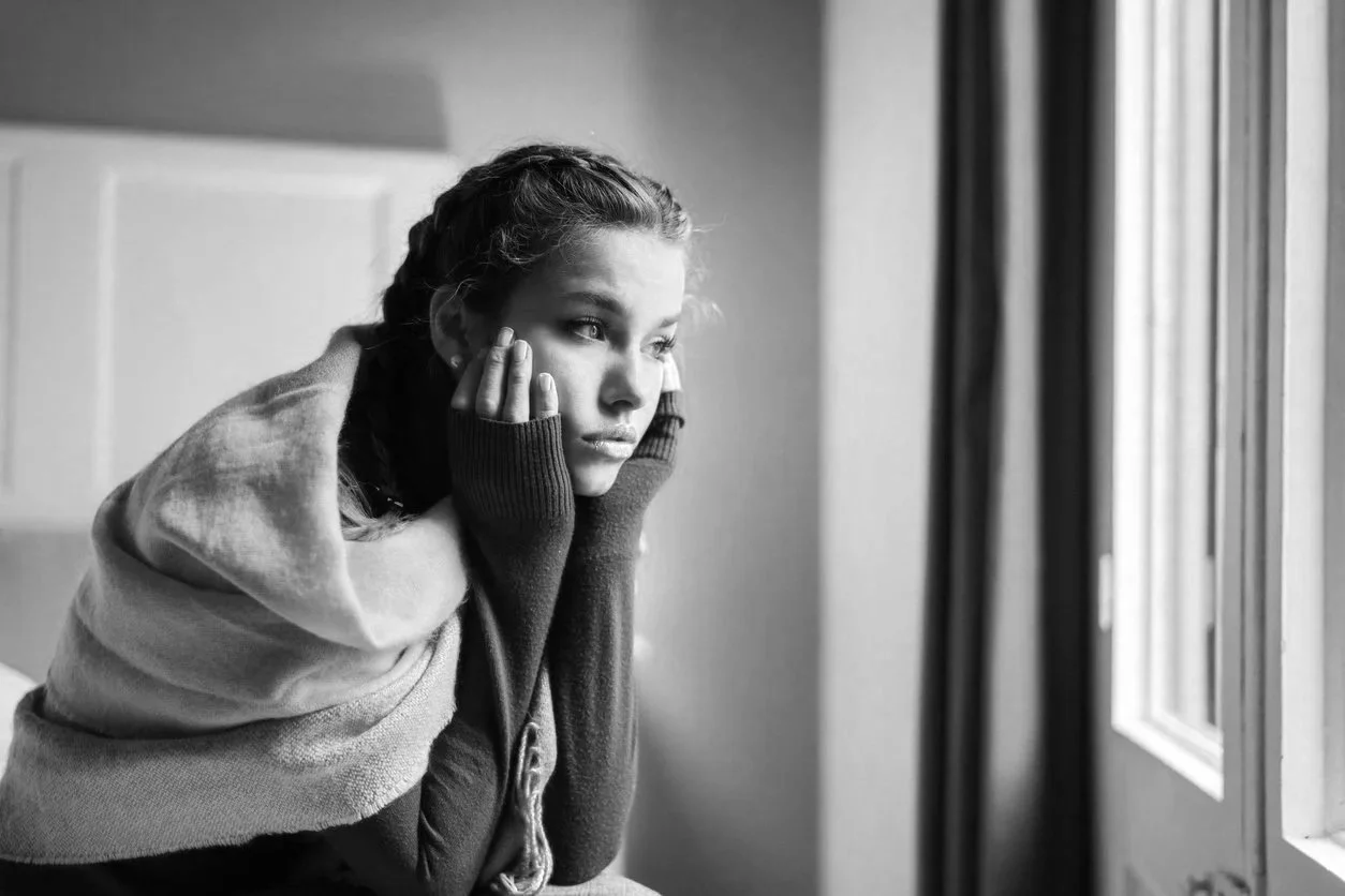 A young woman with braided hair, wearing a hoodie and gloves, looking outside a window with a contemplative expression.