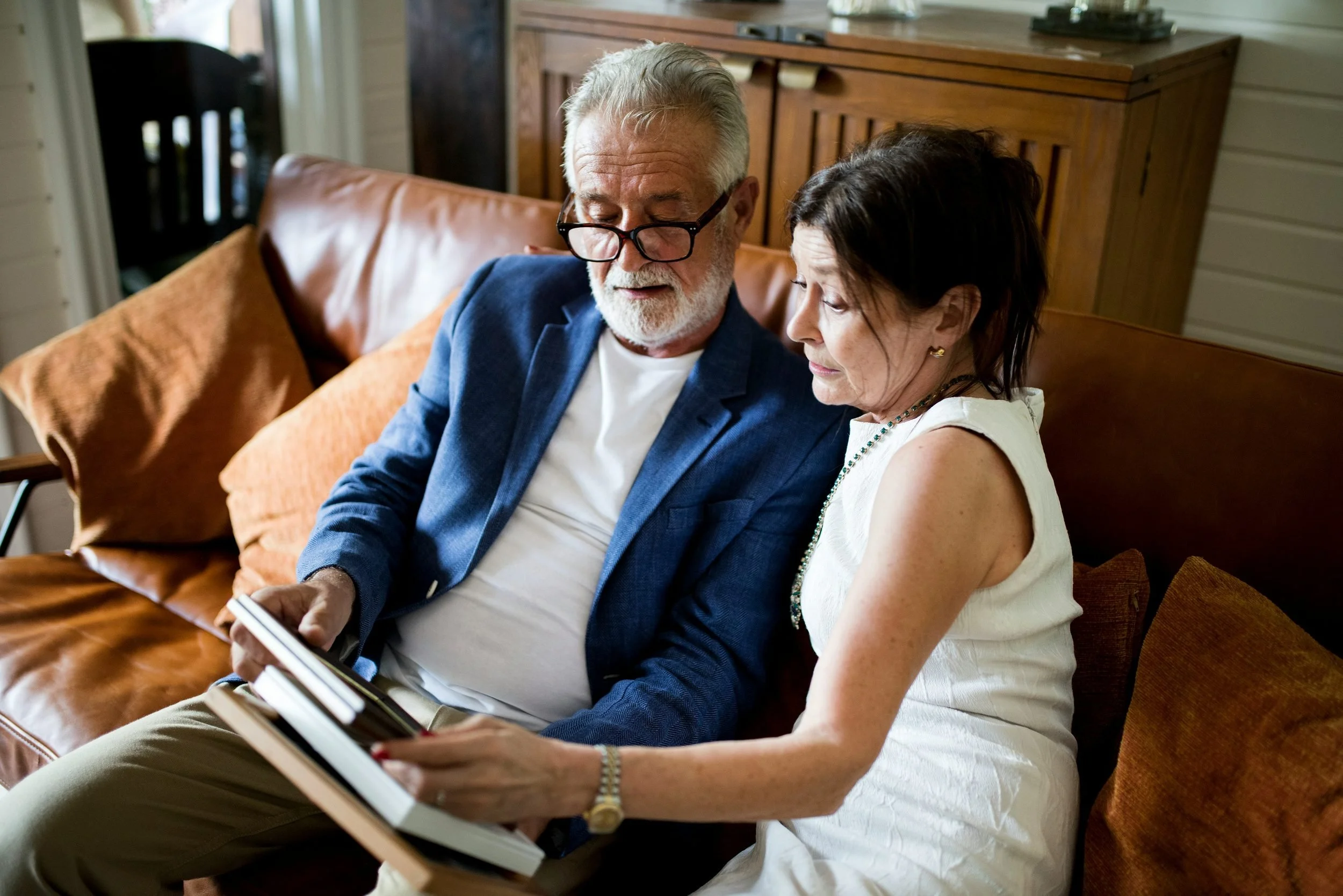 An older man with gray hair, glasses, and a beard wearing a blue blazer, and an older woman with dark hair in a white dress, sitting close together on a brown leather couch, looking at a magazine or book.