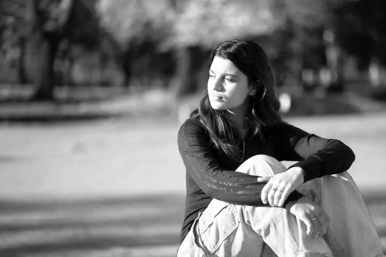Black and white photo of a woman sitting outdoors, looking to the side with a pensive expression, surrounded by blurred trees and grass.