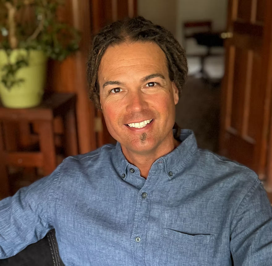 Man with dark curly hair, light skin, and a goatee, wearing a blue button-up shirt, smiling at the camera inside a wooden-paneled room with a potted plant in the background.