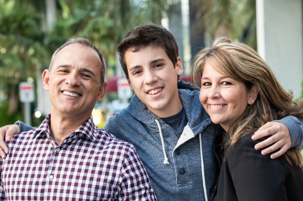 Family of three smiling, hugging outdoors with trees and buildings in the background.