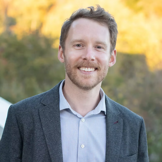 A smiling man with light brown hair and a beard, wearing a gray blazer over a light blue collared shirt, standing outdoors with blurred trees and yellow foliage in the background.