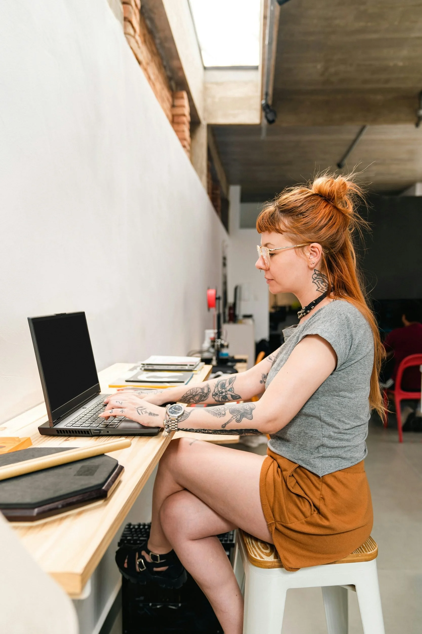A woman with red hair and tattoos is sitting on a high stool, typing on a laptop placed on a wooden desk in a modern workspace with a brick and wooden ceiling. She is wearing glasses, a gray t-shirt, and orange shorts.