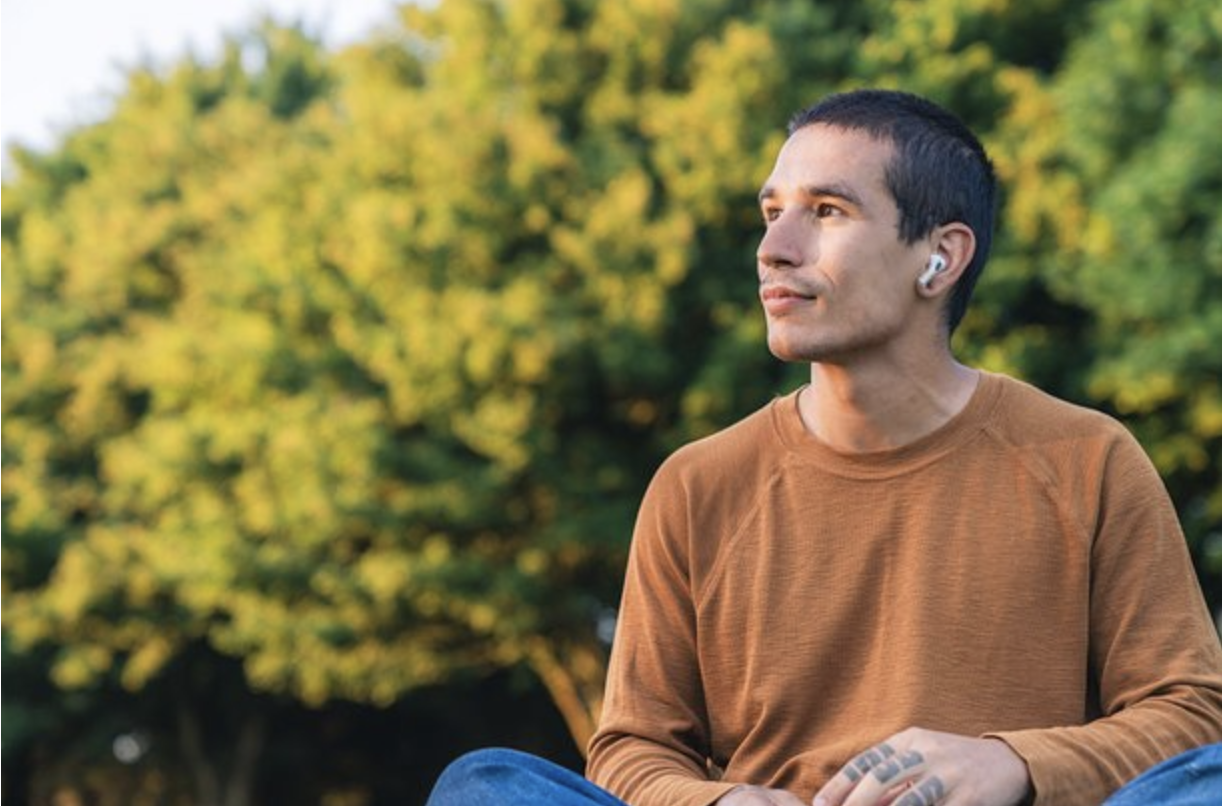 A young man with short dark hair, wearing a brown long-sleeve shirt, sitting outdoors with a blurred green leafy background. He is looking slightly to the side and has white earbuds in his ears.