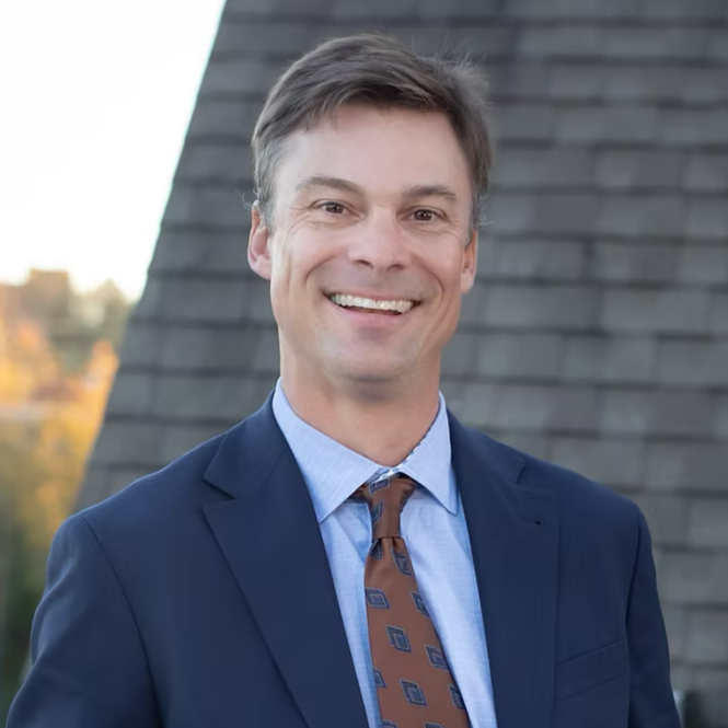A smiling man in a navy blue suit, light blue shirt, and patterned brown tie standing outdoors with a building and trees in the background.