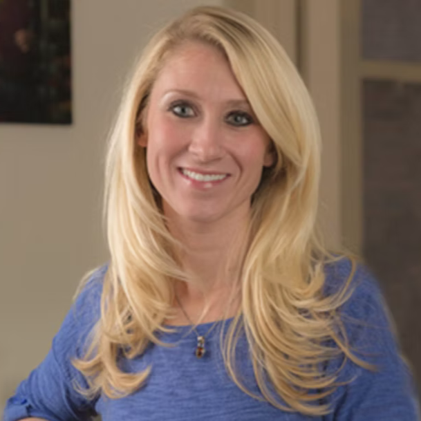 A smiling blonde woman with long hair wearing a blue top, standing indoors against a blurred background.
