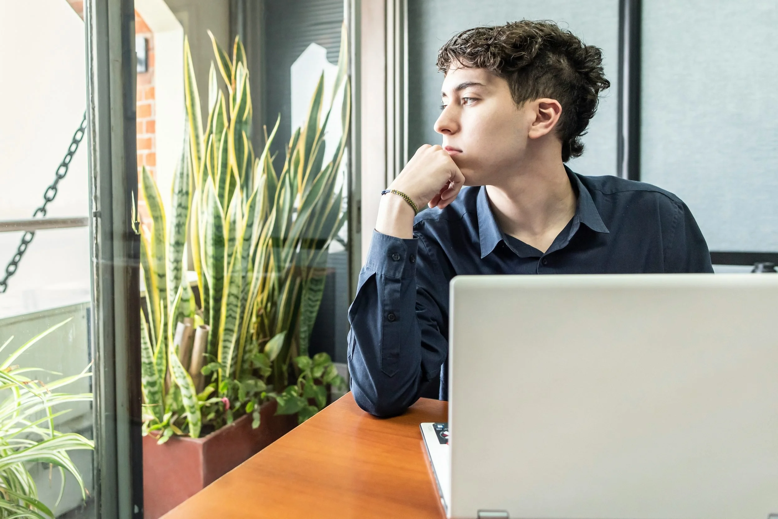 A young man with curly hair and a dark shirt sitting at a wooden table, looking out the window with his hand resting on his chin. There are green plants inside the room near the window, and a laptop is open on the table in front of him.