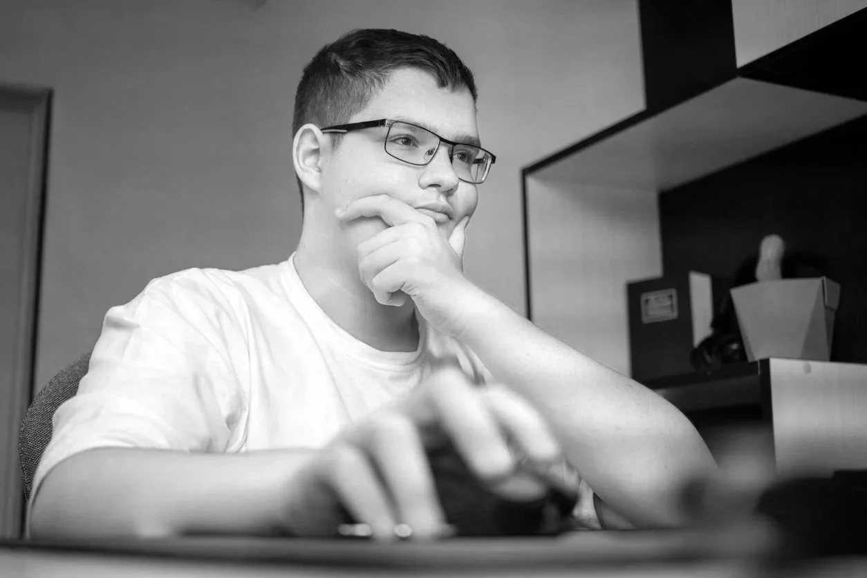 A young man wearing glasses and a white t-shirt sitting at a desk, looking at a computer monitor with a thoughtful expression, one hand on his chin and the other on a mouse.