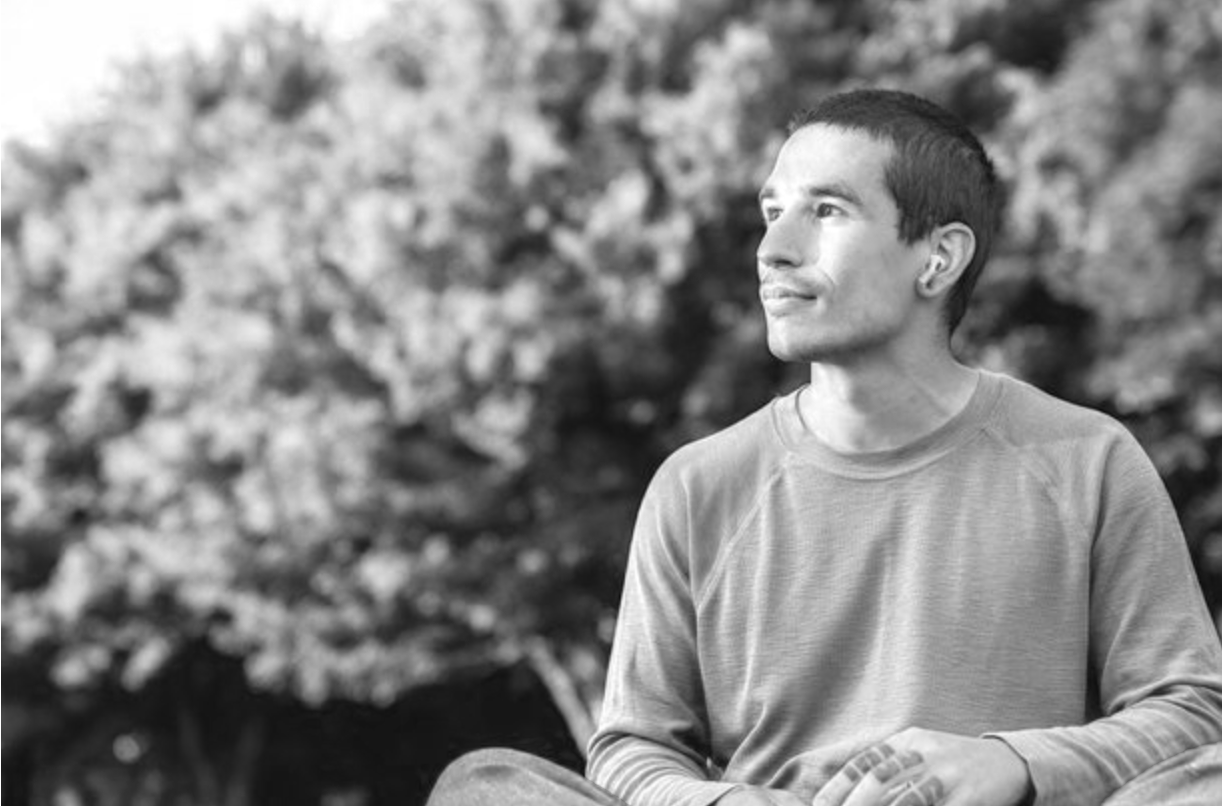 A young man with short hair sits outdoors against a blurred background of trees, looking thoughtfully into the distance.