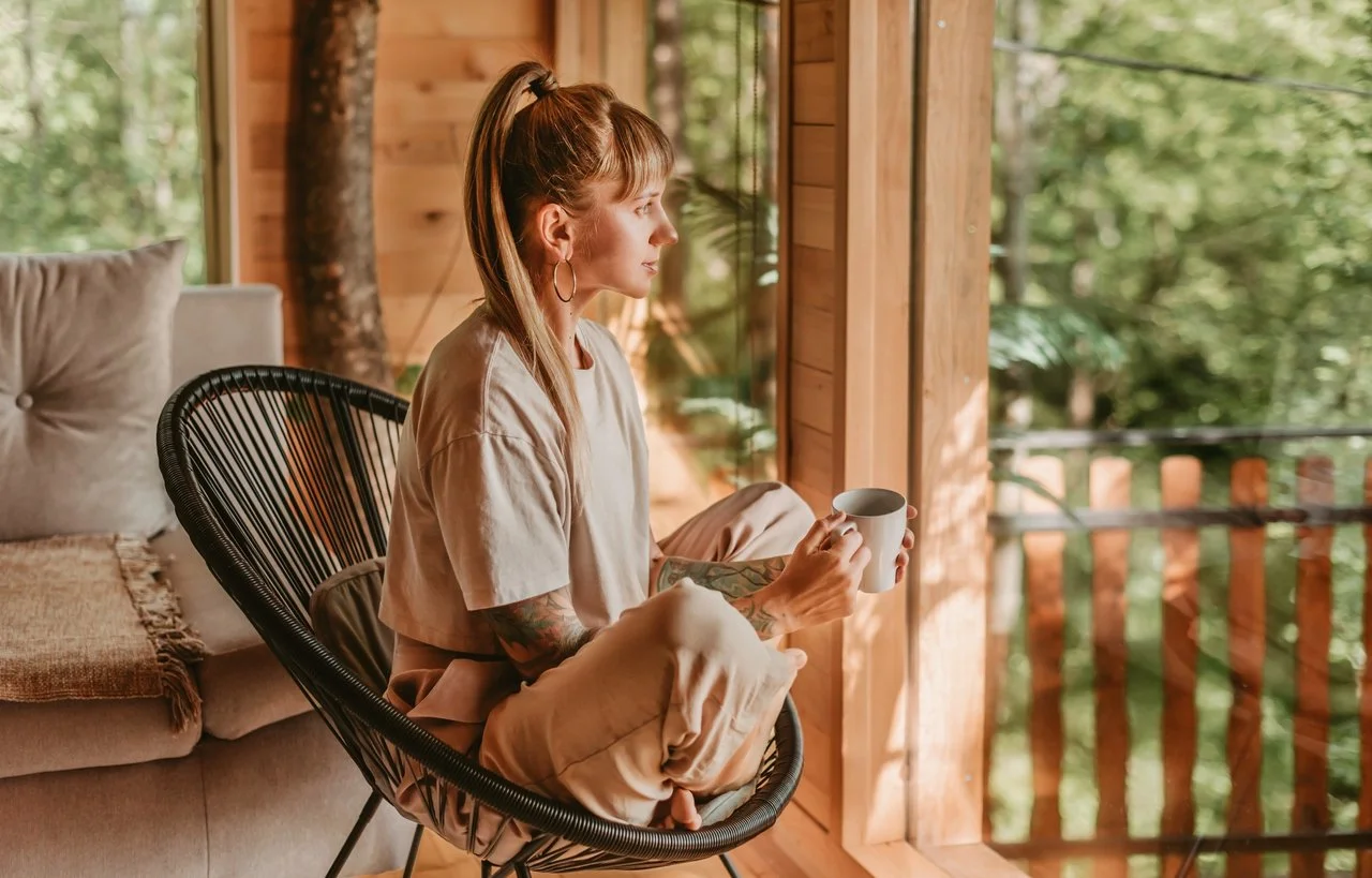 A woman sitting cross-legged in a black outdoor chair, holding a white mug and looking out a glass window onto a wooded area, in a cozy wooden interior.