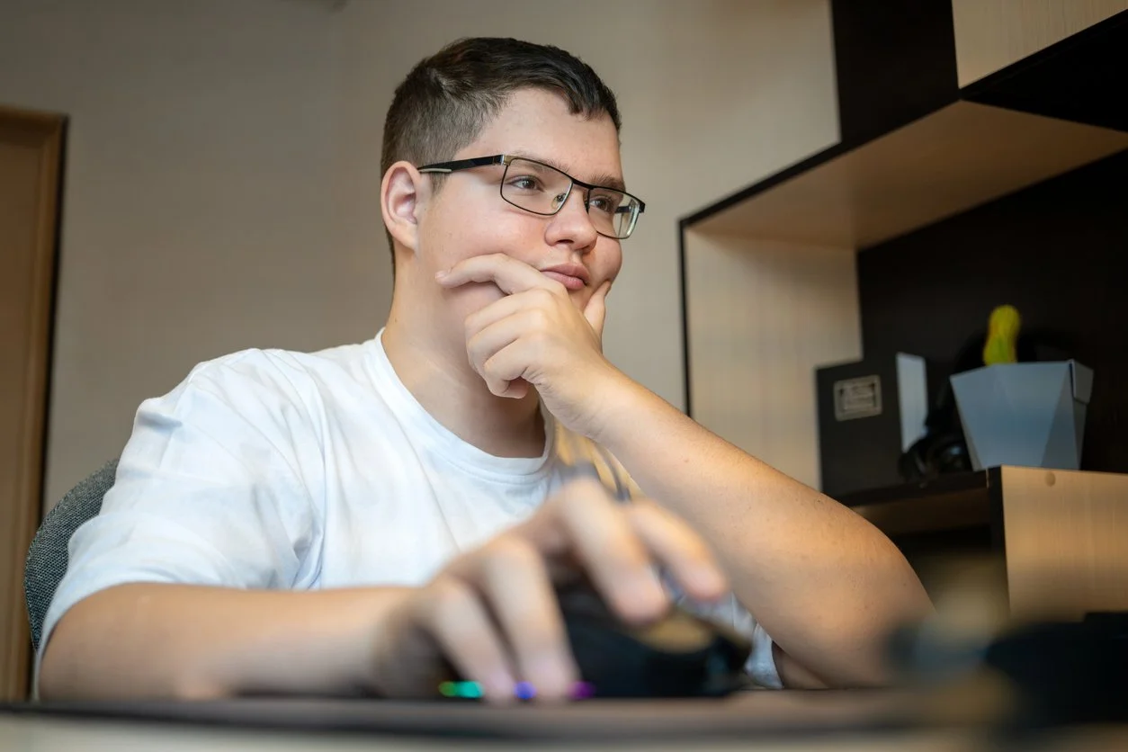 A young man with glasses sits at a desk, watching a computer screen with a thoughtful expression, tapping his chin with one hand and using a mouse with the other.