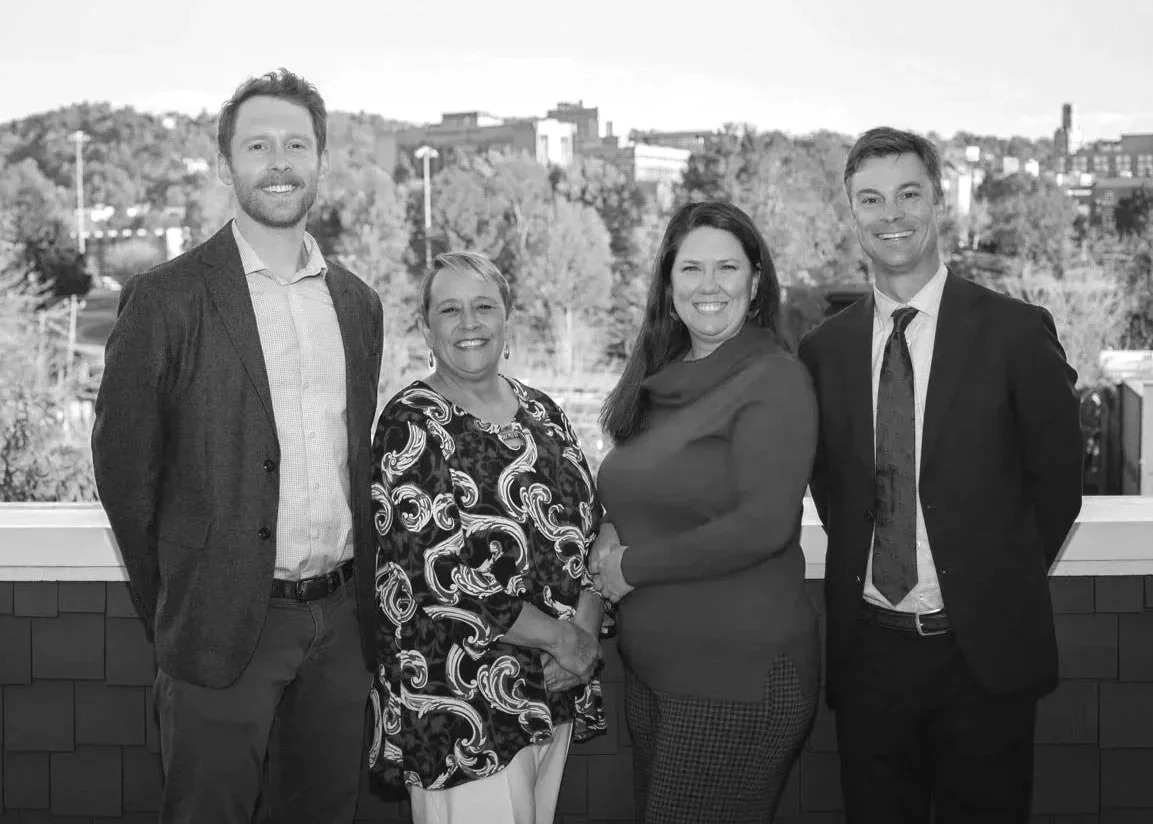 Four professionals, two men and two women, standing outside on a balcony with city buildings and trees in the background, smiling and dressed in business attire.