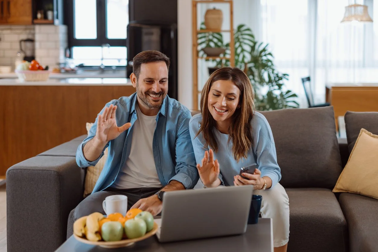 A man and woman sitting on a couch, smiling, with a laptop, microwave, and fruit on the table, in a cozy living room.