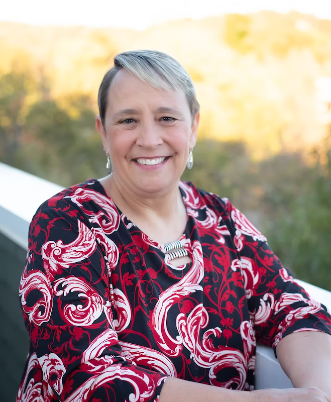 A smiling woman with short blonde hair, wearing earrings, a bold patterned dress with red, white, and black swirls, and a statement necklace, outdoors during what appears to be late afternoon or early evening.