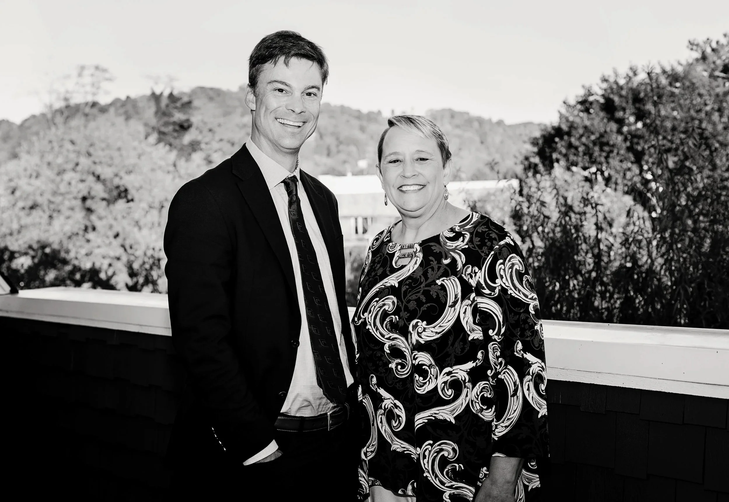 Black and white photo of a man in a suit and a woman in a patterned dress standing outdoors on a balcony, smiling, with a background of trees and hills.