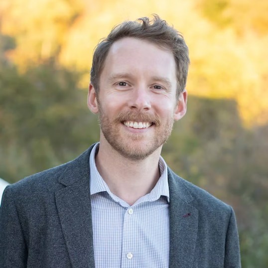 A smiling man with short brown hair and a beard, wearing a blue checkered shirt and gray blazer, standing outdoors with fall foliage in the background.