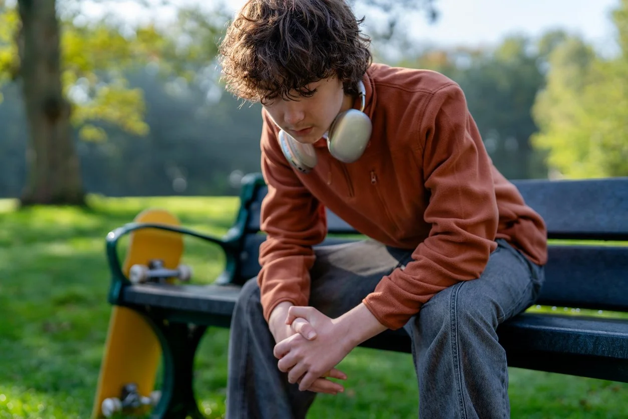 Young man with curly hair sitting on a park bench, wearing headphones, in a contemplative pose with hands clasped, a skateboard resting on the bench beside him, and trees in the background.