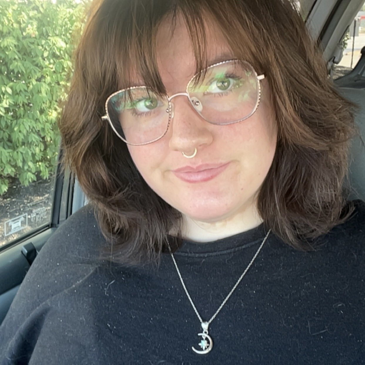 A photo of the artist, a woman with wavy brown hair, glasses, a septum piercing, and a moon and star necklace sitting in a car, with green foliage visible outside the window.