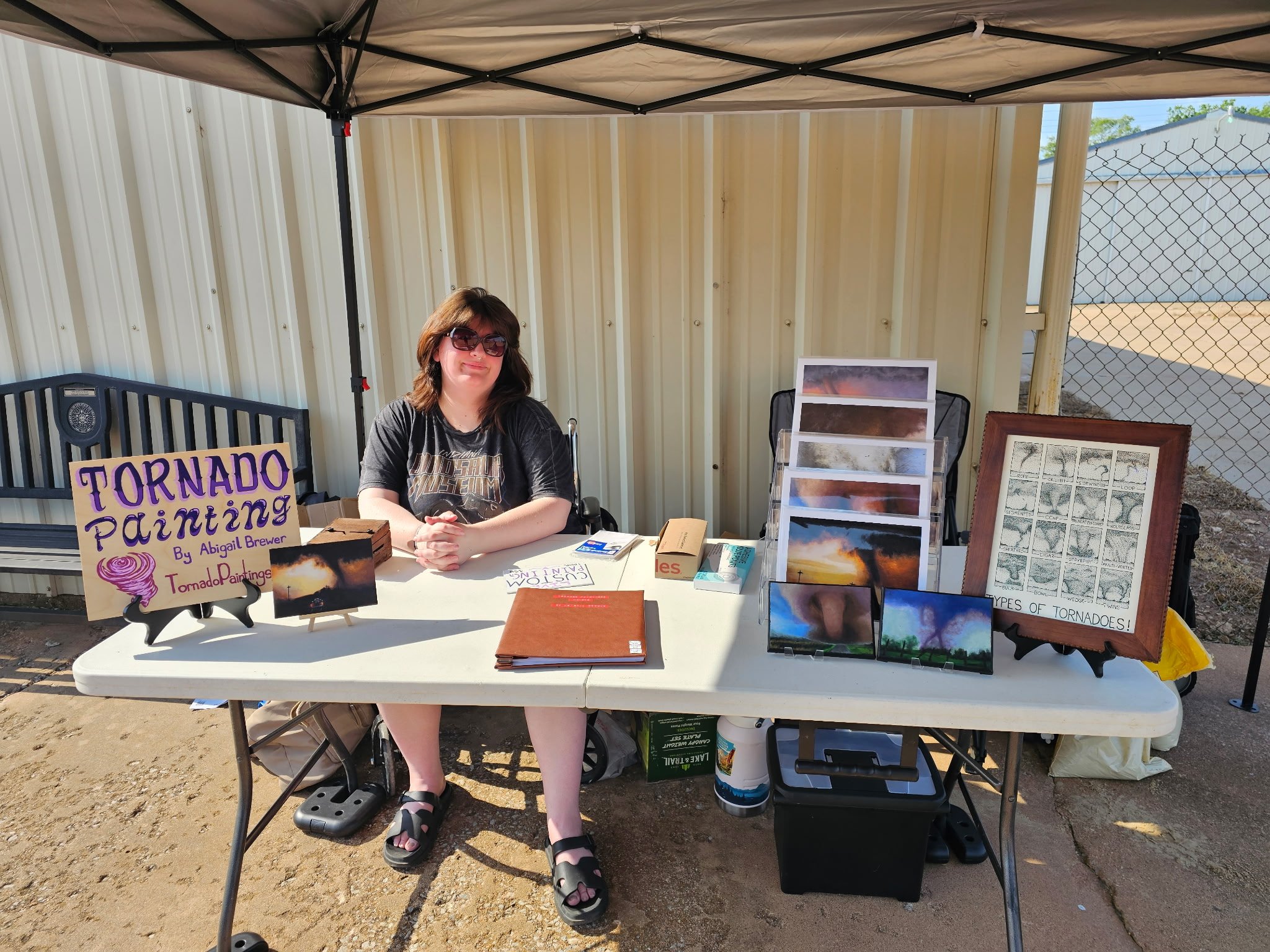 Woman sitting at a booth selling tornado paintings, with signs and pictures of tornado scenes displayed on the table. The booth is under a canopy with a metal fence and a beige building in the background.