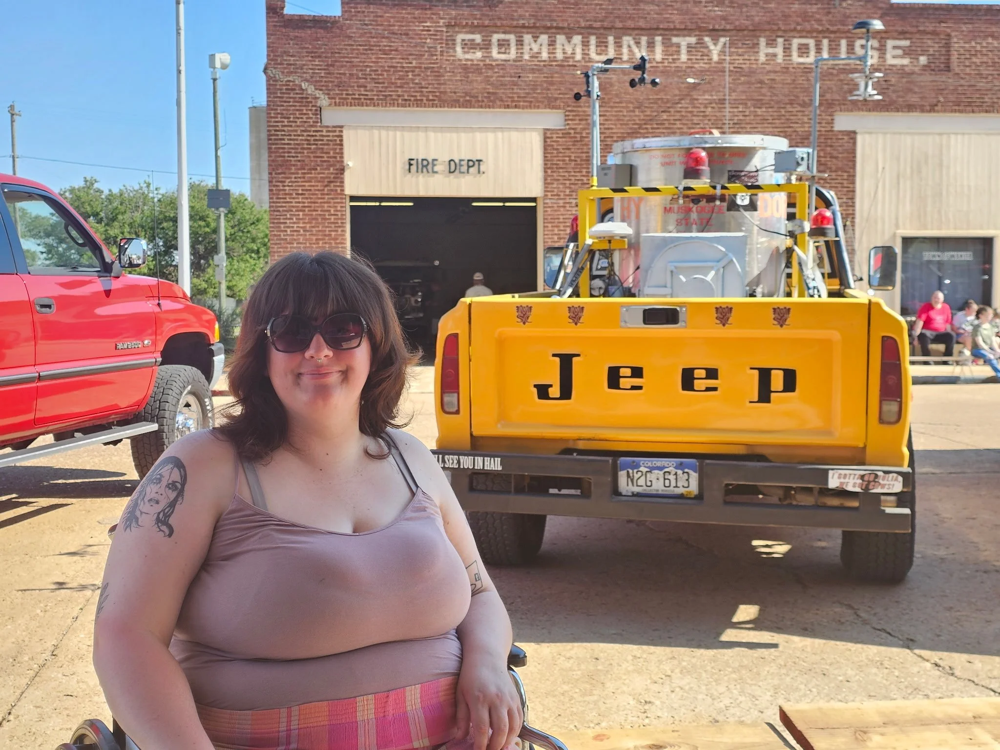 A woman with dark hair and tattoos on her arm, wearing sunglasses and a pink tank top, smiles while standing outdoors in front of a yellow Jeep truck with a large water tank in the back, parked in front of a brick community house with a fire department sign and a garage.