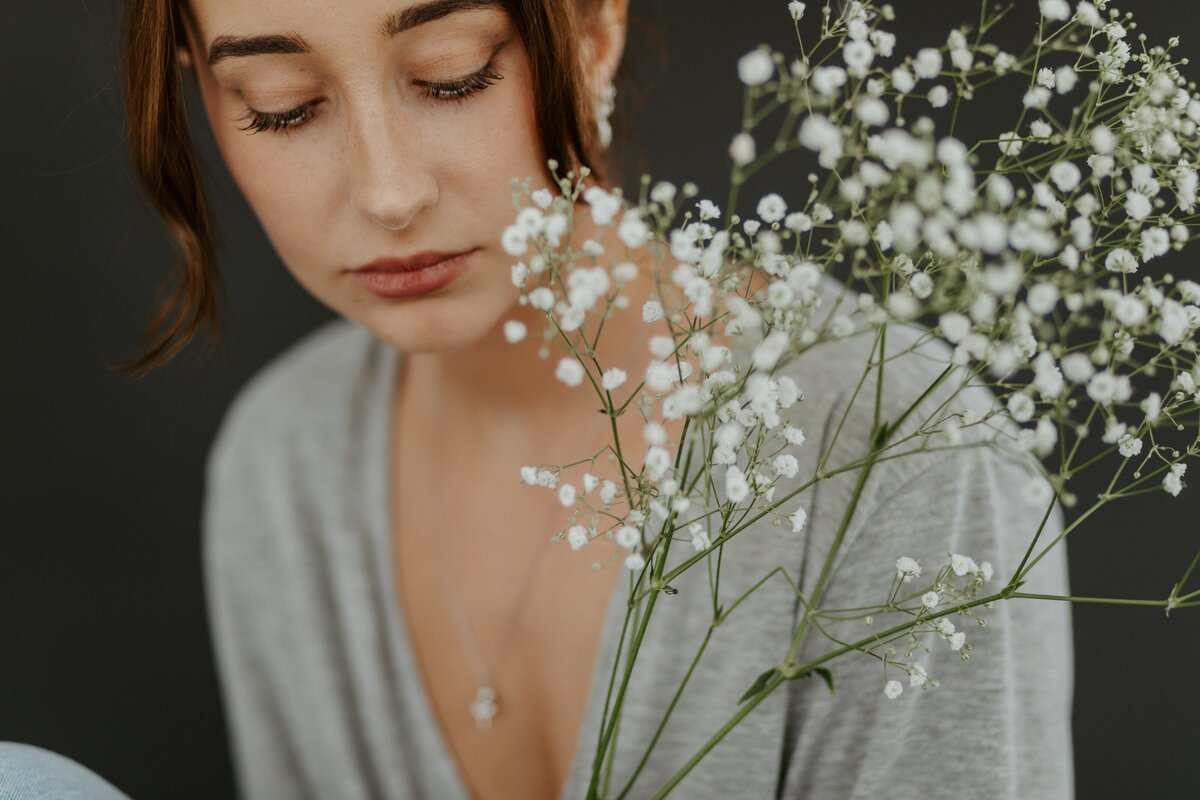A woman with closed eyes and light makeup, wearing a gray top and a delicate necklace, holding a bouquet of baby's breath flowers against a dark background. Thoughtful about women's health, hormone balance, and weight management.