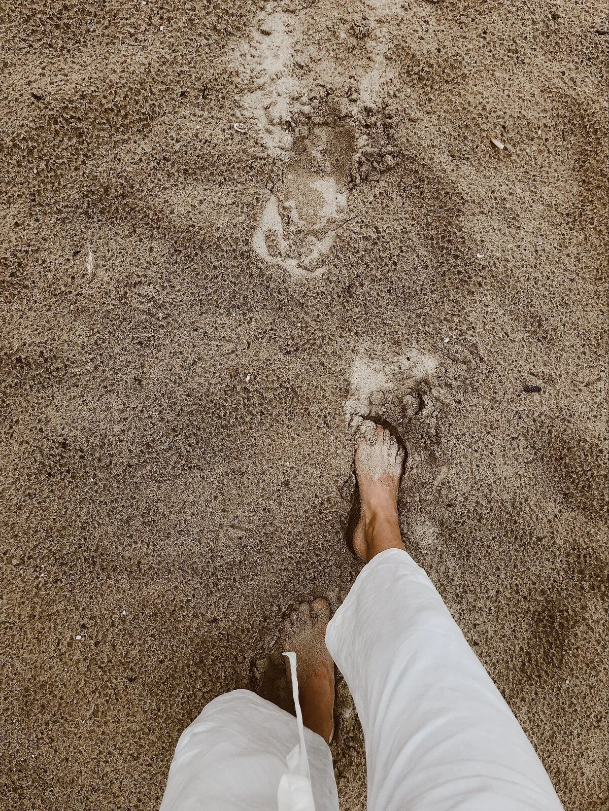 Person walking barefoot on sandy beach, footprints in the sand.